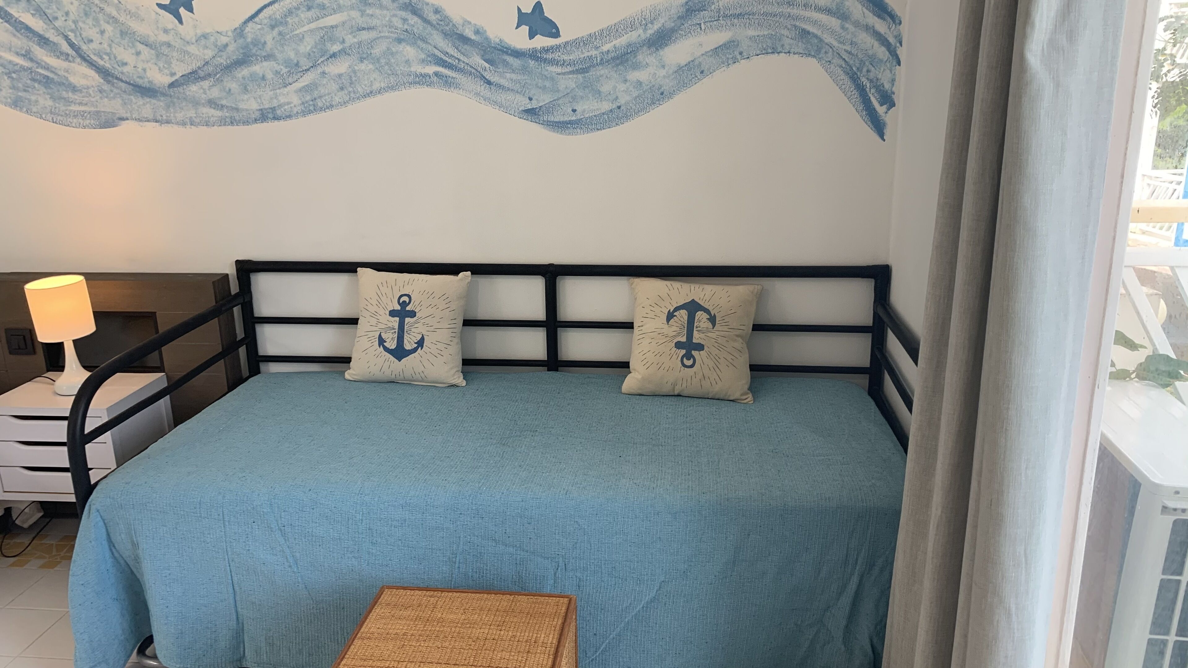 Uma cama com colcha azul e duas almofadas com estampas de âncoras, uma mesa de cabeceira com abajur e um mural na parede com ondas e peixes.