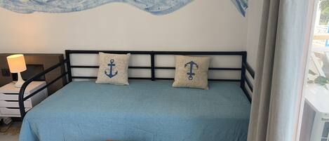 Uma cama com colcha azul e duas almofadas com estampas de âncoras, uma mesa de cabeceira com abajur e um mural na parede com ondas e peixes.