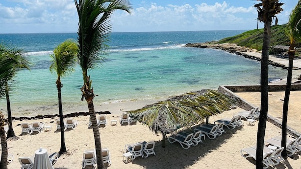 Una playa con sillas y mesas blancas, palmeras y vistas al océano.