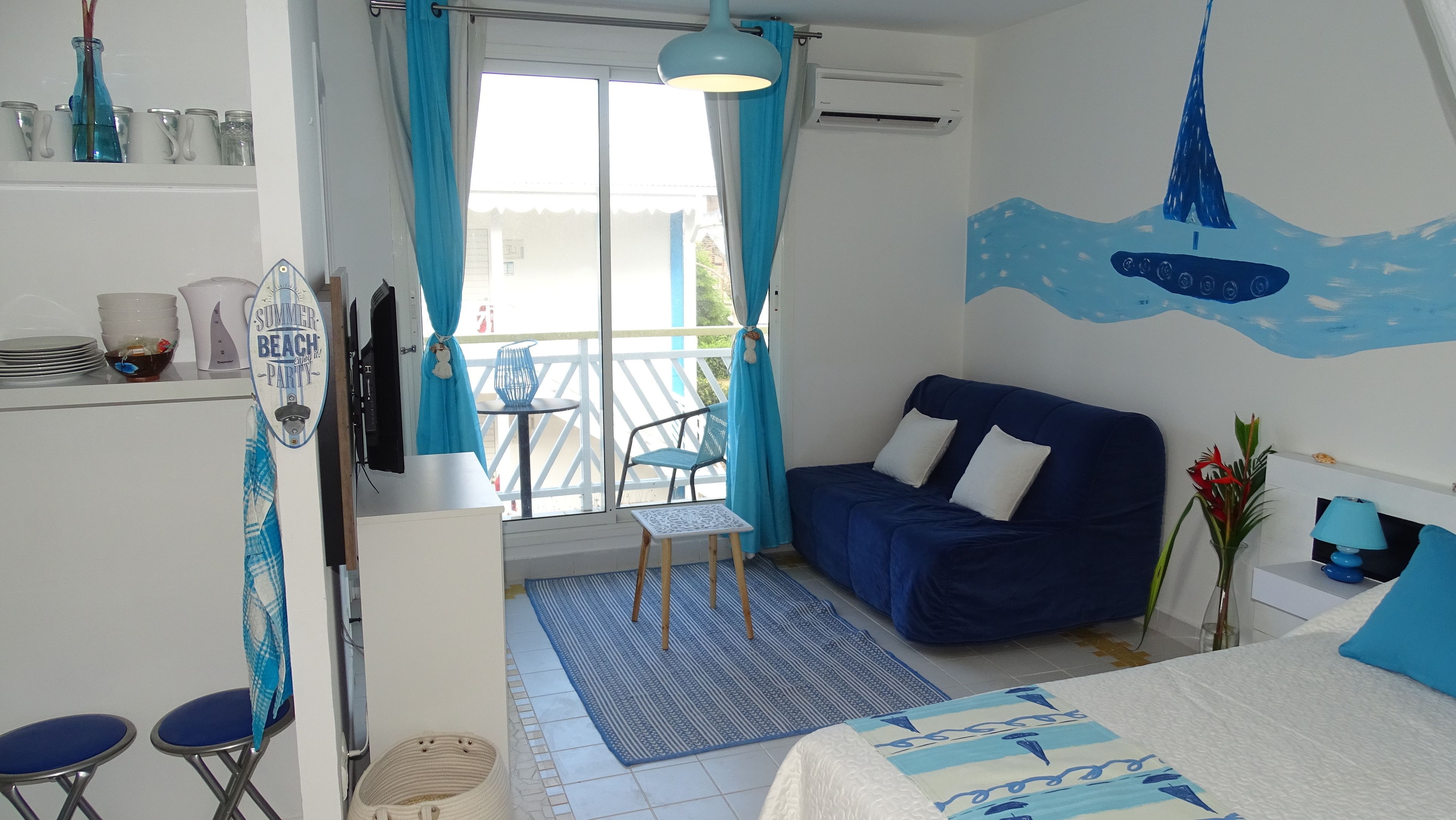 Une pièce comprenant un lit, un canapé bleu, une petite table avec des chaises, une fenêtre avec des rideaux et un mur orné d’une fresque représentant un voilier.