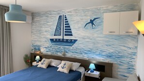 Un dormitorio con cama, mesitas de noche, escritorio, lámpara y un mural en la pared con un velero y una gaviota.