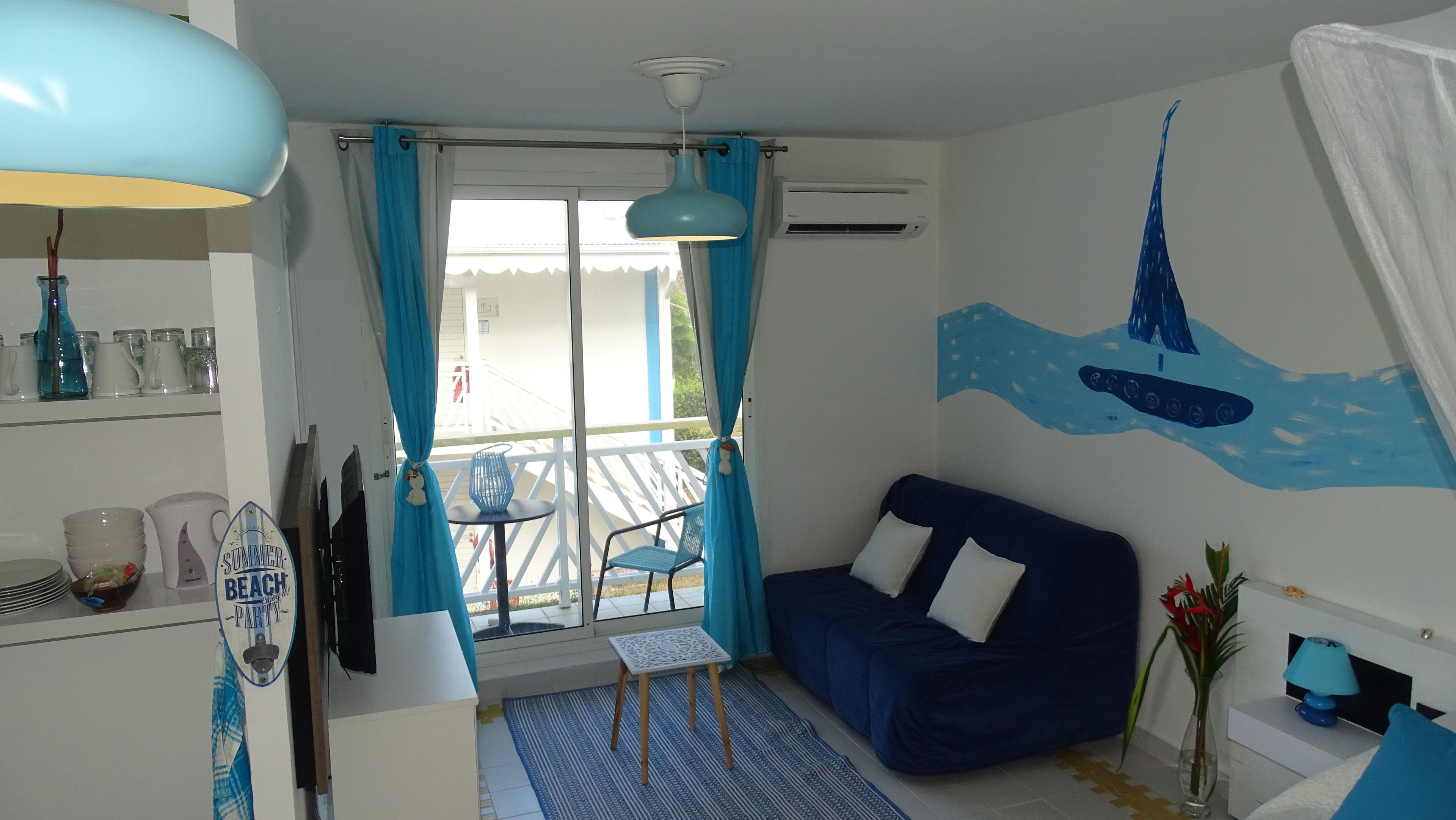 A room with a blue sofa, a small table, a chair, a decorative wall mural, and a balcony with a railing.