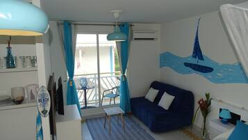 A room with a blue sofa, a small table, a chair, a decorative wall mural, and a balcony with a railing.