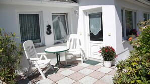 A small white house with a tiled patio, two white chairs, a small table, and a potted plant with red flowers. - Vacation apartment 1 - Ferienanlage Bräuning (Binz)