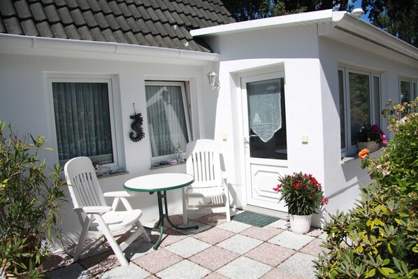 A small white house with a tiled patio, two white chairs, a small table, and a potted plant with red flowers.