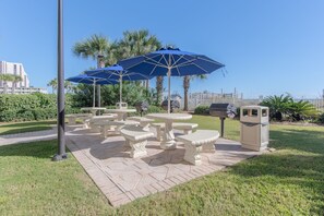 Outdoor seating with tables and benches, umbrellas, and a trash can in a landscaped area with palm trees and a view of buildings.