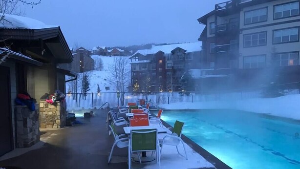 Area piscina esterna con sedie e tavoli, circondata da edifici e un paesaggio innevato.