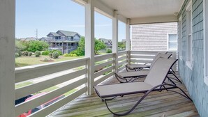 A covered porch with two lounge chairs, a wooden deck, and a view of a house and greenery. - Sandy Bottoms: Oceanside, Pool, Spa, Dogs OK (Nags Head)