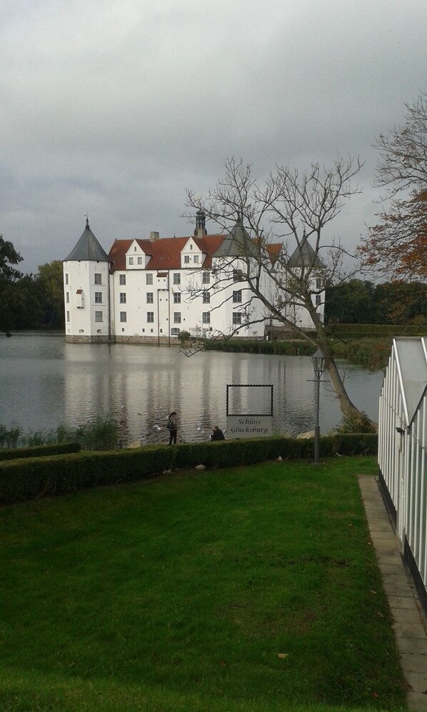 A large, white building with a red roof by a lake, surrounded by greenery.