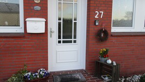 Una casa de ladrillo rojo con puerta blanca, número 27, un buzón, una guirnalda y una pequeña mesa con flores.