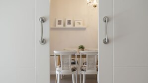 Un coin repas avec une table et des chaises blanches, une petite plante en pot et une étagère murale ornée de tableaux encadrés.