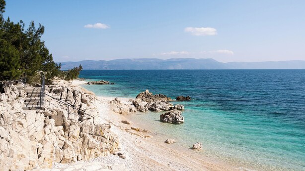 A rocky beach with clear turquoise water, a staircase leading down to the shore, and distant mountains under a clear sky. - Holiday apartment with Interntet access (Rabac)