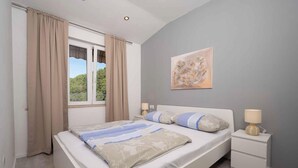 A bedroom with a bed, a nightstand, a lamp, a painting, and a window with curtains. - Holiday apartment with Interntet access (Rabac)