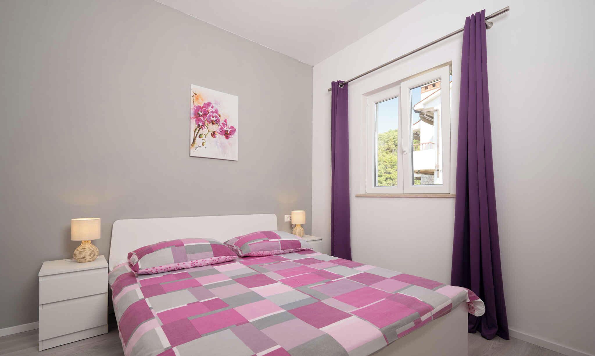 A bedroom with a bed, two bedside tables, a window with purple curtains, and a wall-mounted picture.