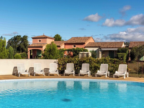 A swimming pool with lounge chairs, a building with a tiled roof, and greenery in the background.
