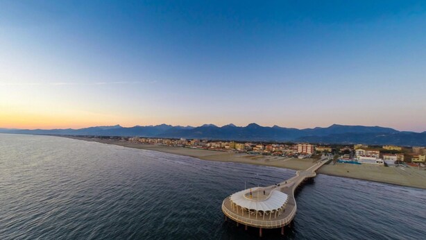 Vista aérea de una zona costera con un edificio circular distintivo cerca del agua, una playa y una cadena montañosa al fondo.