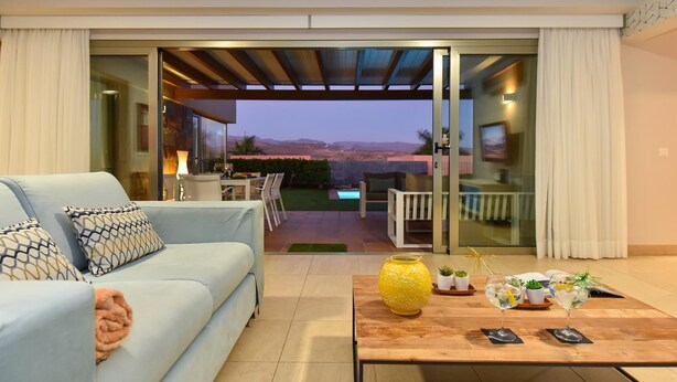 A modern living room with a sofa, a coffee table with decorative items, and a sliding glass door leading to an outdoor patio with a pool and seating area. - Par 4 Rental 9 Salobre by VillaGranCanaria (Maspalomas)
