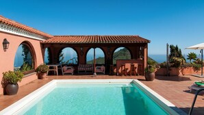 Ein Poolbereich mit freiem Blick auf das Meer, ein überdachter Sitzbereich mit Stühlen und Tischen sowie eine Dachkonstruktion mit Fliesen.