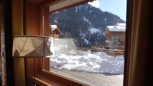 Ein Fenster mit Blick auf eine verschneite Berglandschaft, eine Holzhütte und verschneite Erde.
