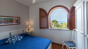 A bedroom with a bed, a chair, a window with a view, a ceiling fan, and a framed picture on the wall. - 1 - House by the sea with pool near Cefalù (Campofelice di Roccella)