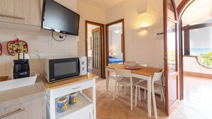 A compact kitchen with a microwave, coffee maker, and a TV mounted on the wall. There is a dining area with a table and chairs, and a balcony with a view. - 1 - House by the sea with pool near Cefalù (Campofelice di Roccella)