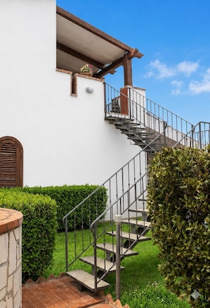 Um edifício branco com uma escadaria que leva a uma varanda, rodeado por vegetação.