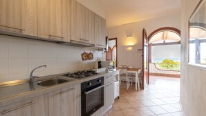 A modern kitchen with wooden cabinets, a stainless steel sink, and a gas stove. There is a dining area with a table and chairs, and a view of the sea through an open archway. - 1 - House by the sea with pool near Cefalù (Campofelice di Roccella)