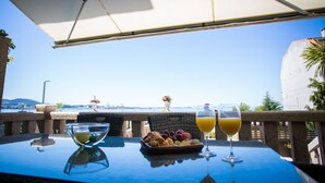 Una mesa con una cesta de fruta, un vaso de zumo de naranja y una copa de vino blanco, bajo una sombrilla, con vistas al mar y edificios al fondo.