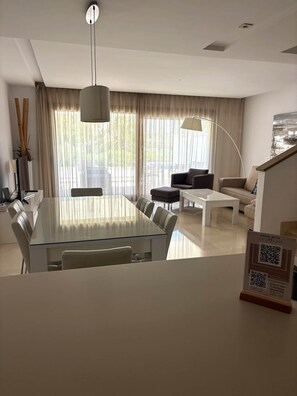 A modern living room with a glass dining table, sofas, and a large window with curtains.