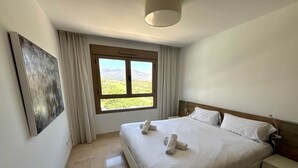 Una habitación de hotel con cama, mesita de noche, lámpara, ventana con vistas y un cuadro en la pared.