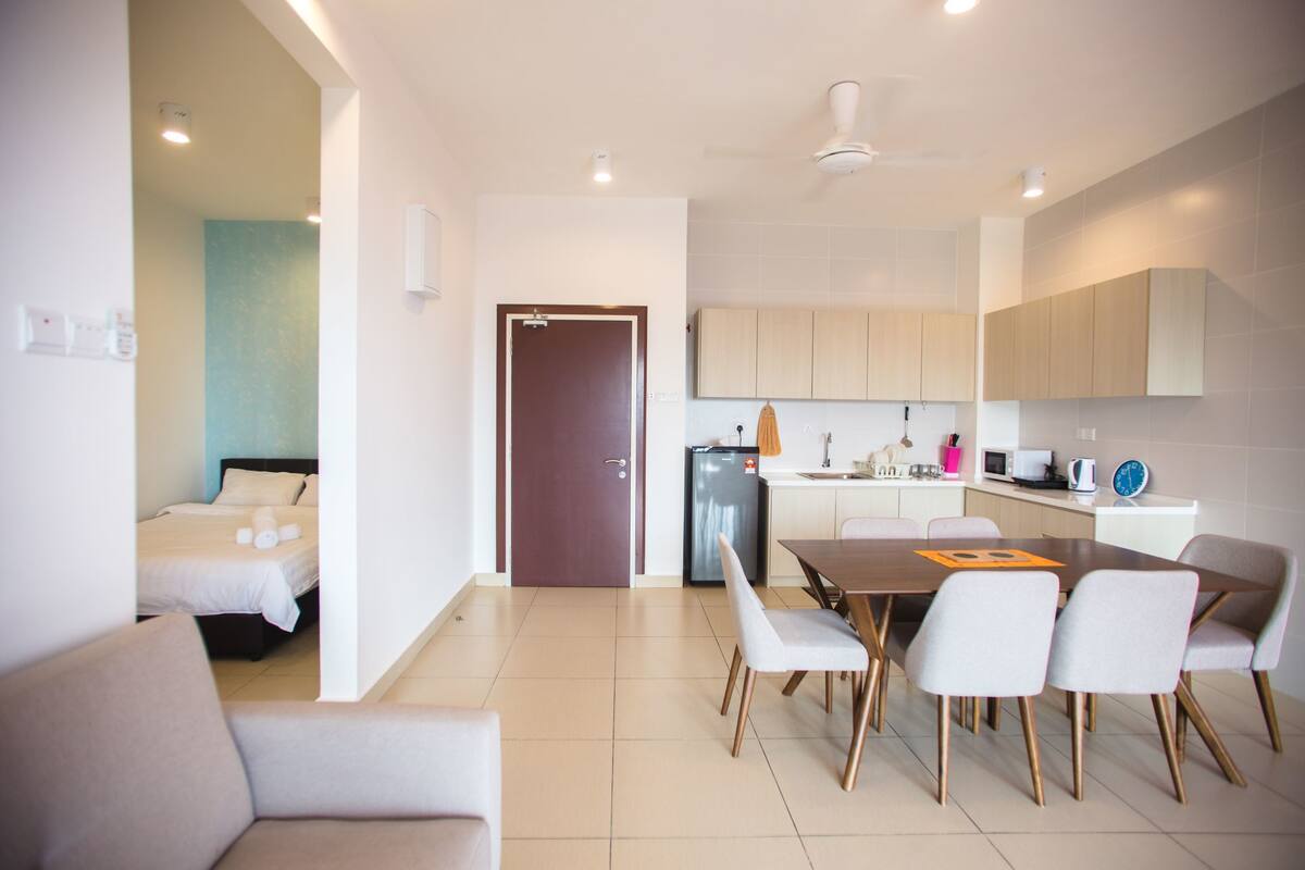 A modern hotel room with a bed, a dining area with a table and chairs, a kitchen area with cabinets and appliances, and a door leading to another room.