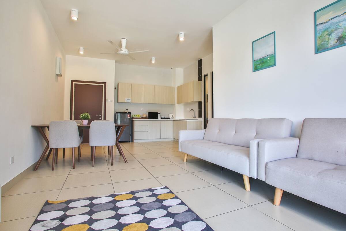 A modern living room with a sofa, dining table, and chairs. A kitchen is visible in the background with cabinets and appliances.