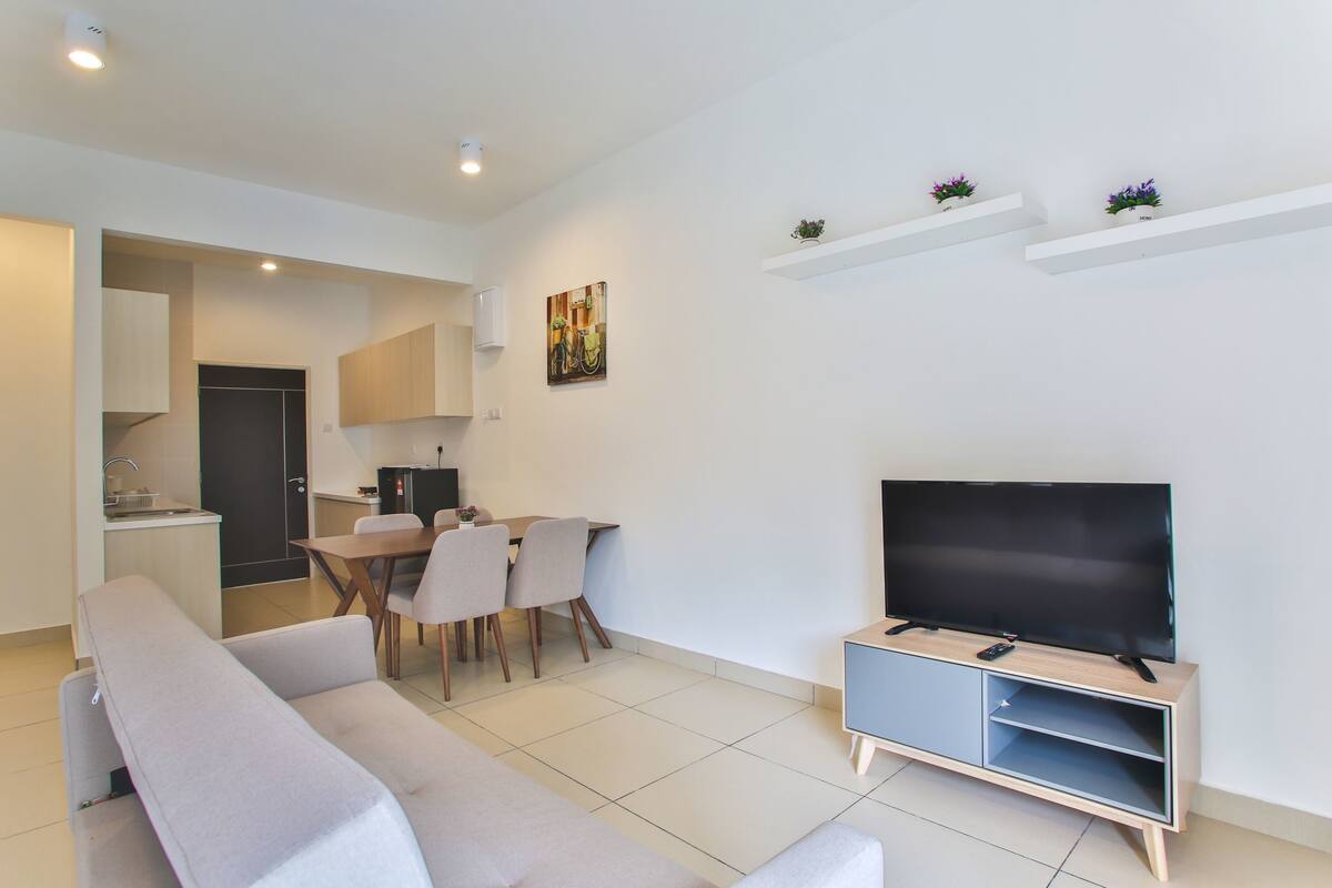 A modern living room with a sofa, dining table, chairs, a TV on a wooden stand, and a kitchen in the background.