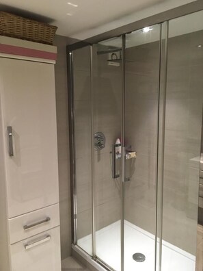 Eine gläserne Duschkabine mit weiß gefliestem Boden, einer chromfarbenen Duschbrause und einem kleinen Ablagefach für Toilettenartikel.