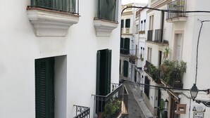 A narrow street lined with white buildings, each with green shutters and balconies adorned with potted plants. - Discover Sitges, steps from the beach. 3 BRM + 2 BATH (Sitges)