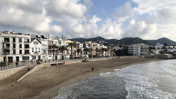 A coastal town with a sandy beach, a promenade lined with buildings, and a mountain in the background. - Discover Sitges, steps from the beach. 3 BRM + 2 BATH (Sitges)