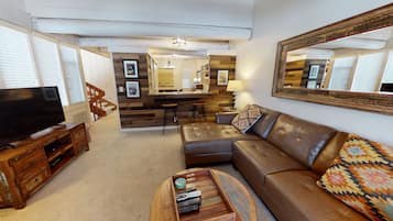 A living room with a brown leather sofa, a wooden coffee table with remote controls, a TV on a wooden stand, a kitchen area with a wooden countertop and bar stools, and a large mirror on the wall.