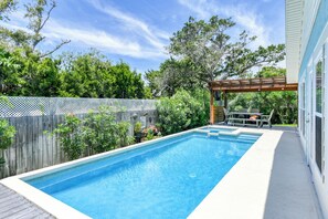 A rectangular swimming pool with a wooden deck surrounding it, a pergola with seating, and a white fence with climbing plants.