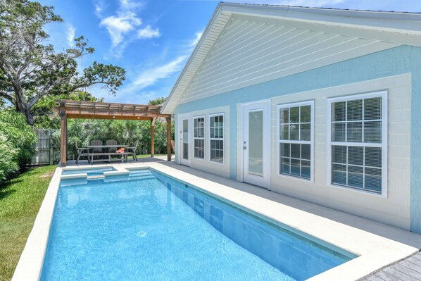 A swimming pool with a pergola, a covered patio, and a house with large windows and a door.