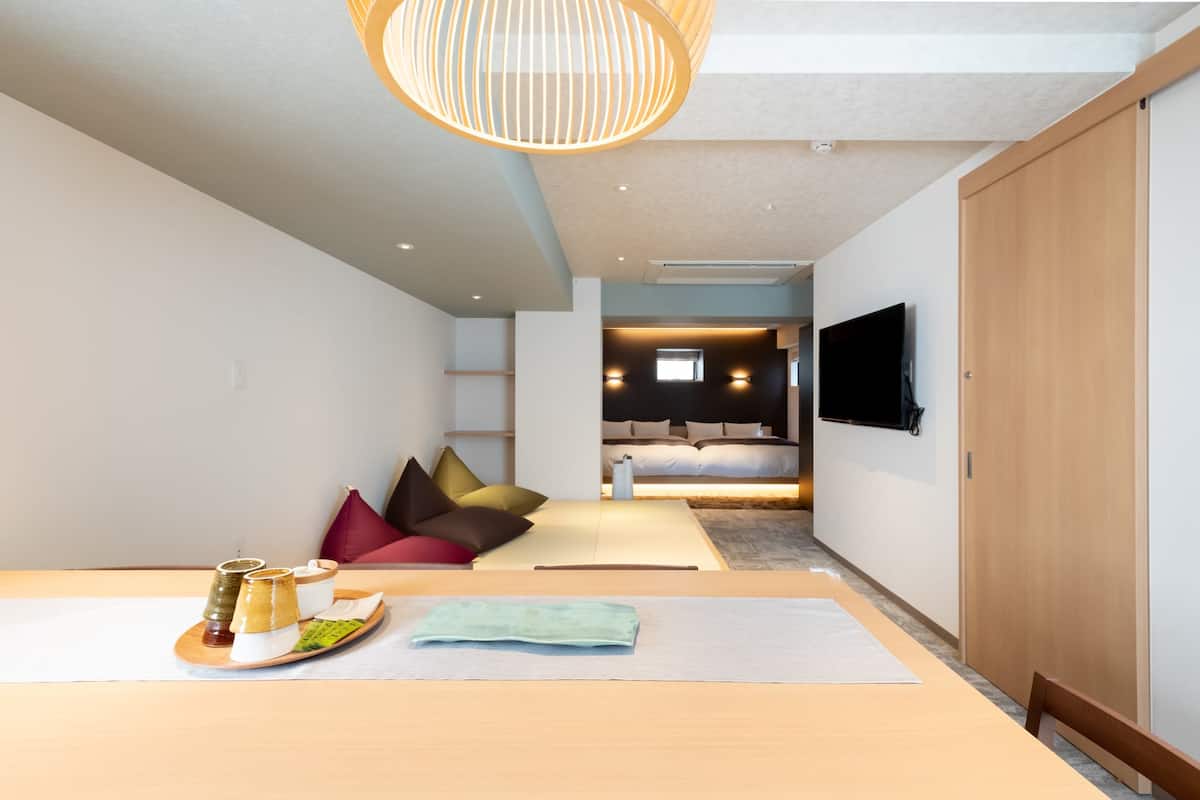 A modern hotel room with a wooden table, a sofa with cushions, a TV mounted on the wall, and a sliding door.
