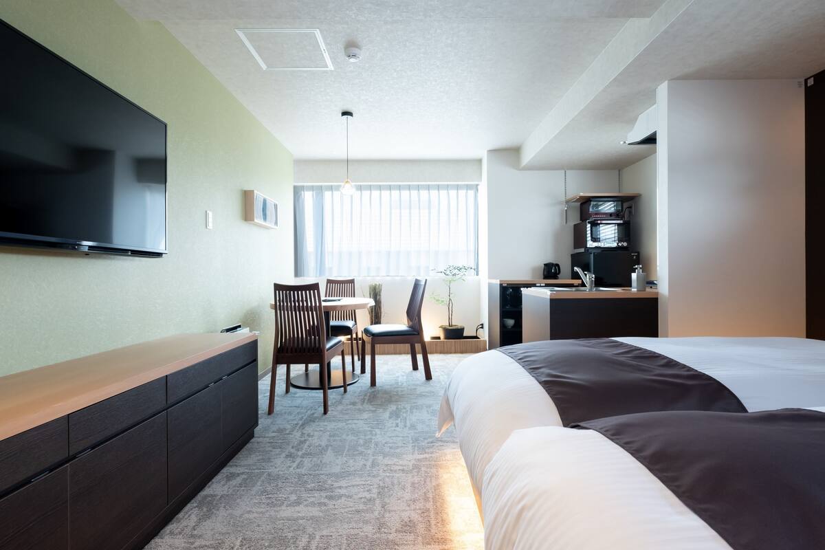 A hotel room with a bed, a desk with a computer, a TV mounted on the wall, and a window with blinds.