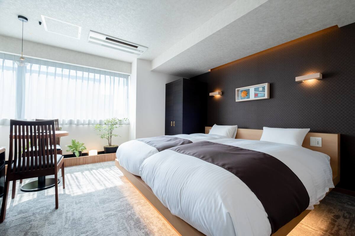 A modern hotel room with a large bed, a wooden chair, a potted plant, and a large window.