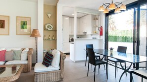 Een moderne woonkamer met een bank, een eettafel met stoelen, een keukenhoek en een groot raam met uitzicht op groen.