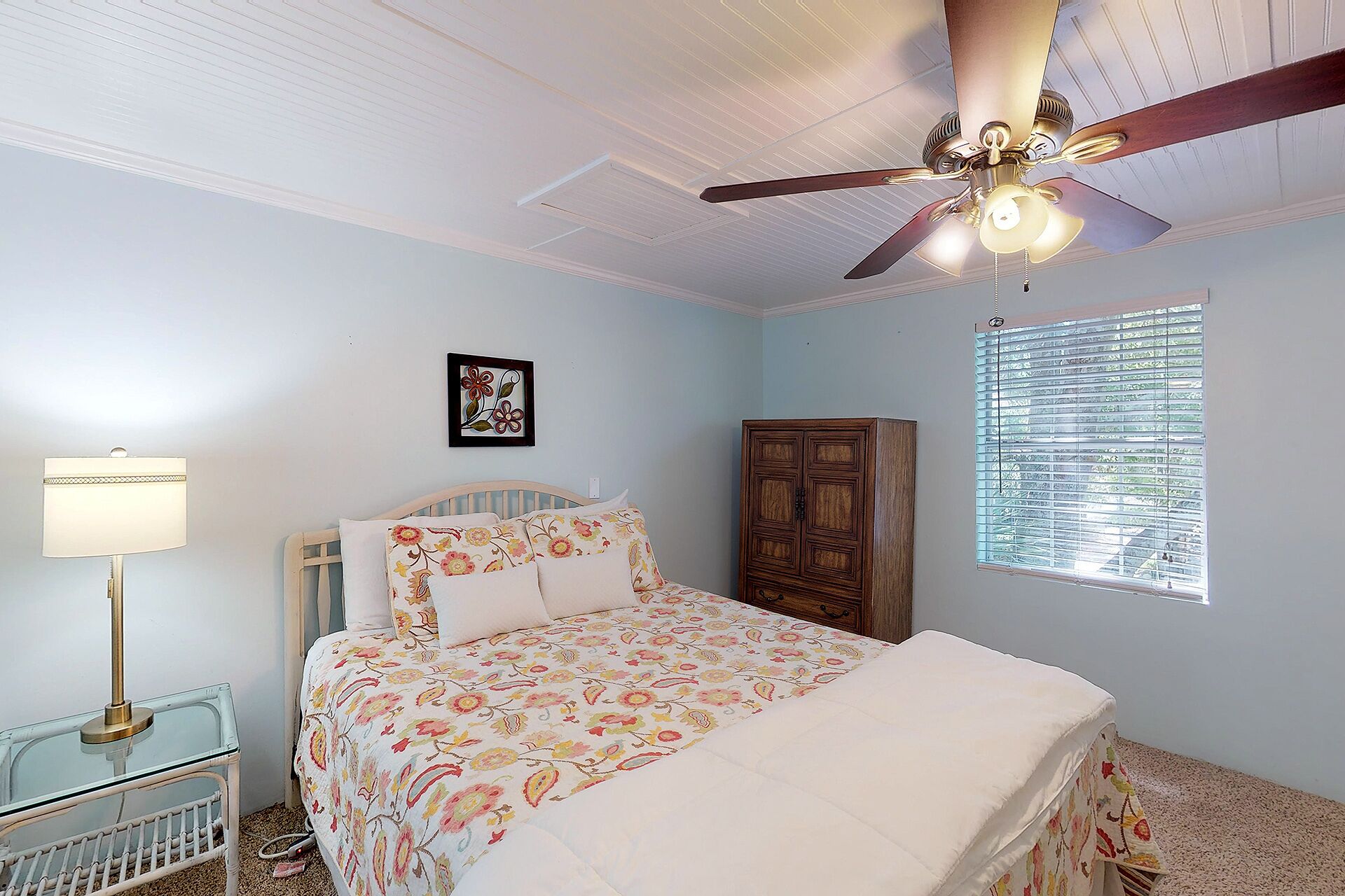 A bedroom with a bed, a ceiling fan, a nightstand, a lamp, a wardrobe, and a window with blinds.