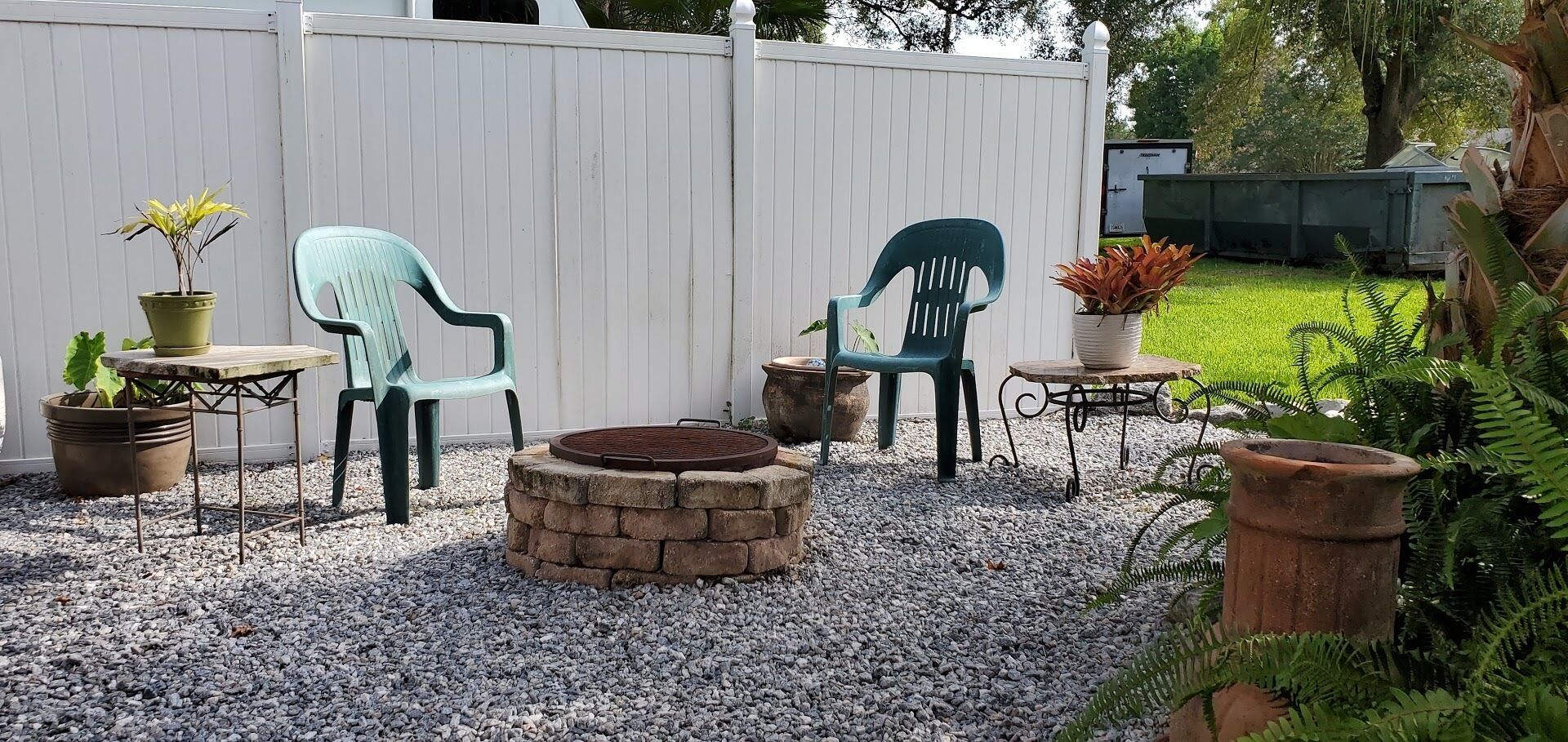 Een patio met twee groene plastic stoelen, een klein rond tafeltje en een vuurkorf. Achtergrond met plantenpotten en een wit hek.