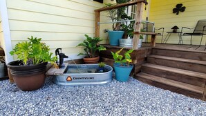 Un petit espace jardin aménagé avec différentes plantes en pot, un élément décoratif aquatique et des marches en bois menant à un porche.
