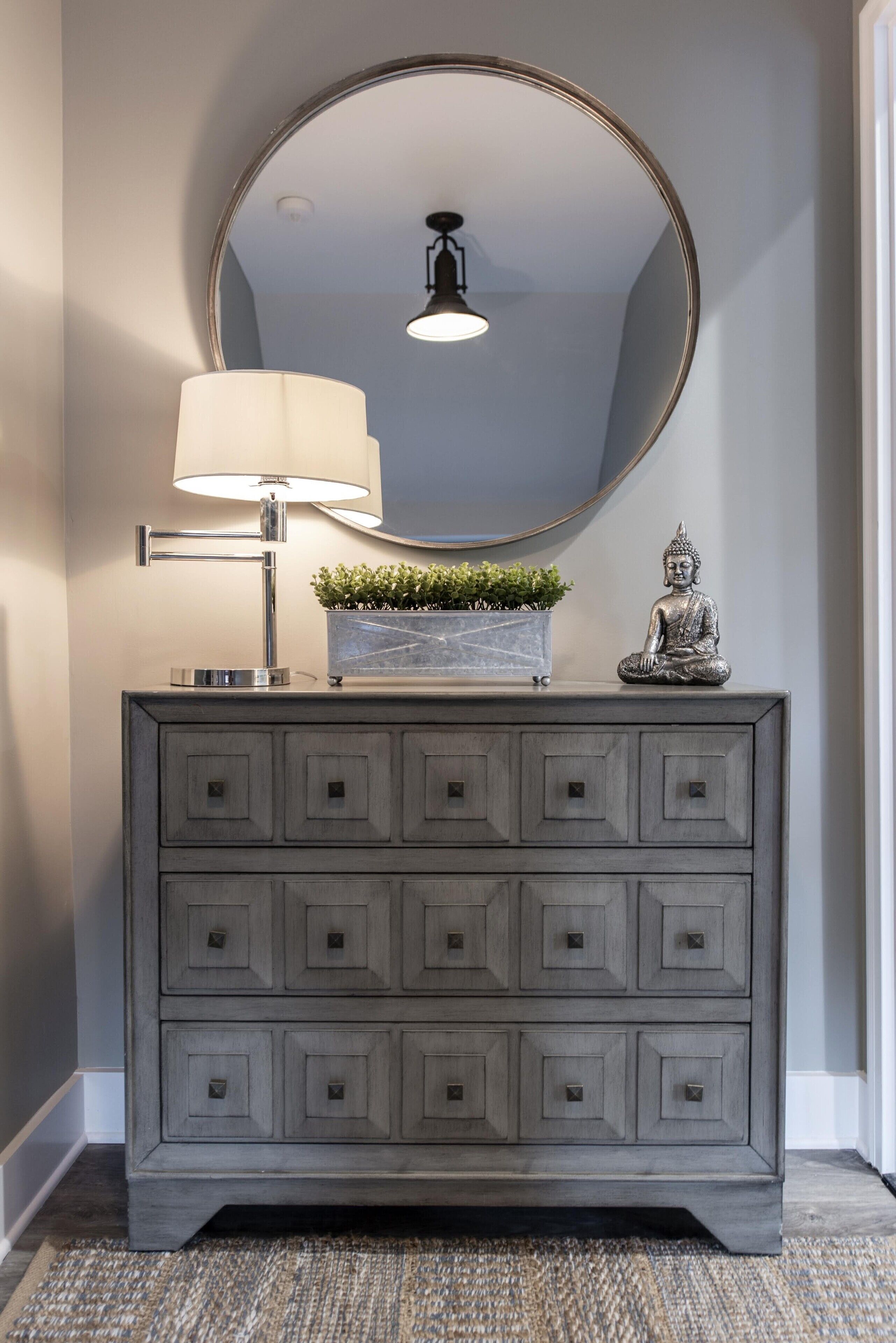A grey dresser with a round mirror, a lamp, and a Buddha statue.