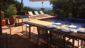 An outdoor pool area with lounge chairs, a dining table set for two, and a view of the ocean. - You cook, eat and relax around the pool with nice sea view. (Palafrugell)