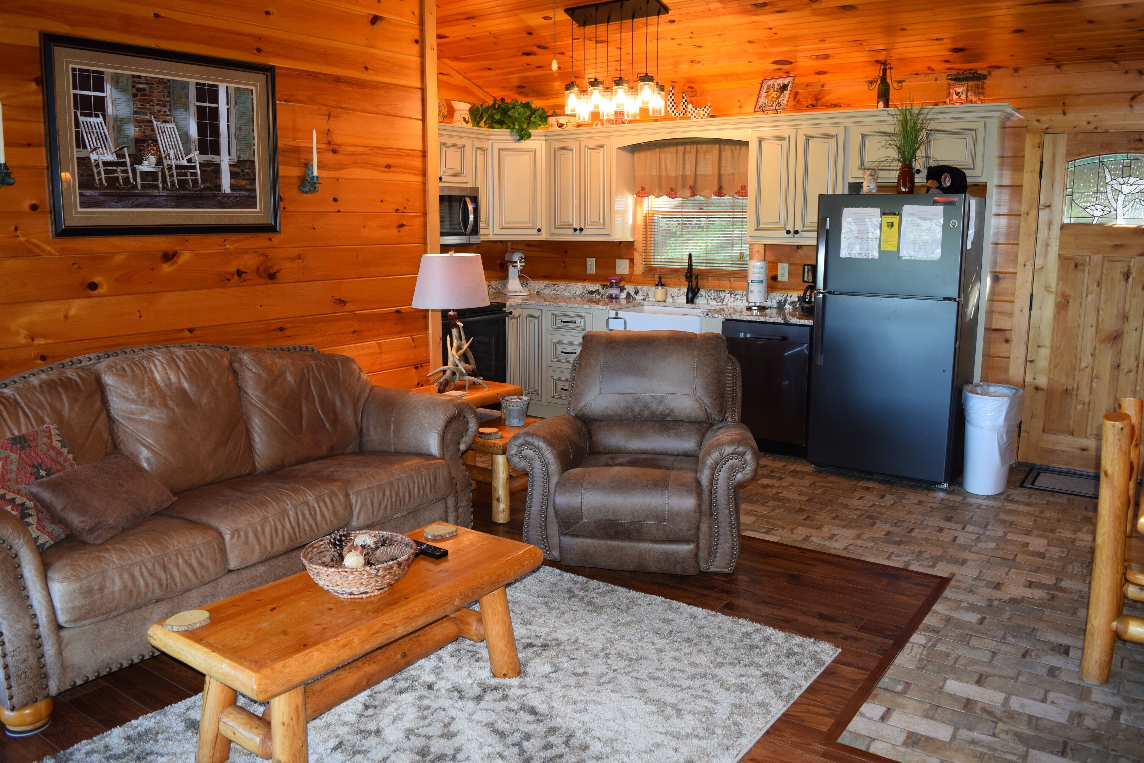 A cozy cabin interior with a leather sofa, a wooden coffee table, a leather armchair, and a kitchen area with wooden cabinets and a refrigerator.