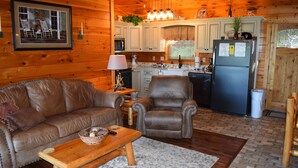 L’intérieur chaleureux d’un chalet comprend un canapé en cuir, une table basse en bois, un fauteuil en cuir et un coin cuisine avec des placards en bois et un réfrigérateur.
